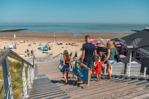 Famiglia scende le scale verso una spiaggia soleggiata, vicino a una lodge sul mare con ombrelloni colorati.