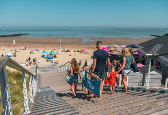 Une famille descend l’escalier vers une plage ensoleillée, proche d’un lodge de plage et de parasols colorés.