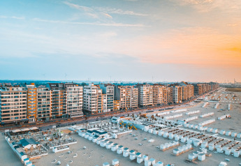 Vue panoramique sur une promenade de plage moderne avec immeubles et cabines blanches au coucher du soleil.