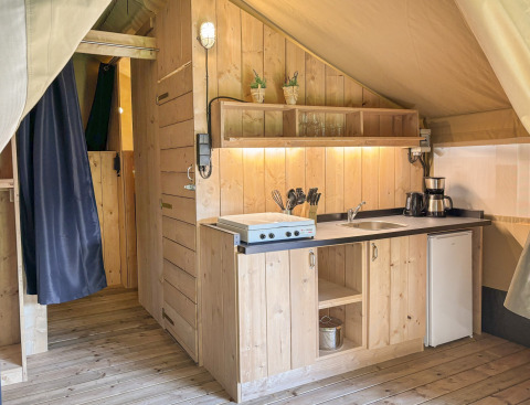 Intérieur d’une tente safari avec cuisine en bois, évier, plaque de cuisson et mini-frigo à Les Rives de Courtes.
