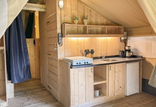 Intérieur d’une tente safari avec cuisine en bois, évier, plaque de cuisson et mini-frigo à Les Rives de Courtes.