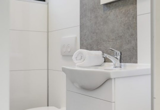 Moderne og kompakt badeværelse med hvidt toilet, håndvask og sammenrullet håndklæde på håndvasken.