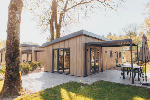 Modern glamping cabin with wooden exterior, covered patio and outdoor furniture in a sunny garden setting.