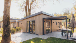 Modern glamping cabin with wooden exterior, covered patio and outdoor furniture in a sunny garden setting.