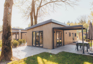 Moderne glamping lodge met houten gevel, overdekt terras en tuinmeubilair in een groene, zonnige omgeving.