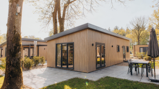 Modern wooden lodge at Loevestein with large windows, outdoor patio, and garden furniture in the sun.