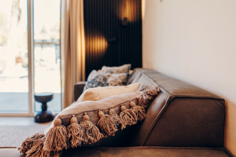 Salon moderne de glamping avec canapé marron, coussin à pompons et lumière naturelle à travers les baies vitrées.