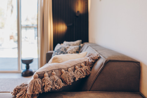 Salon moderne de glamping avec canapé marron, coussin à pompons et lumière naturelle à travers les baies vitrées.