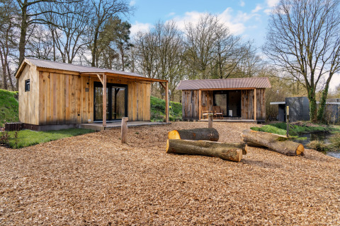 Two wooden cabins with porches in a forest setting, logs and wood chips on the ground under blue sky.