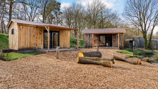 Two wooden cabins with porches in a forest setting, logs and wood chips on the ground under blue sky.