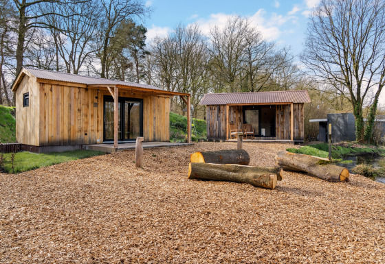 Two wooden cabins with porches in a forest setting, logs and wood chips on the ground under blue sky.