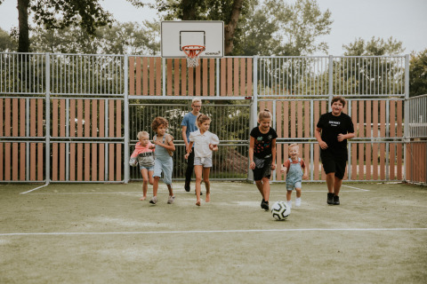 Kinder spielen Fußball auf einem Außenplatz bei einer Glamping-Unterkunft mit Bäumen im Hintergrund.