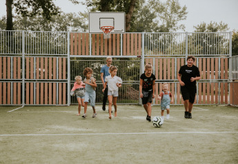 Kinder spielen Fußball auf einem Außenplatz bei einer Glamping-Unterkunft mit Bäumen im Hintergrund.