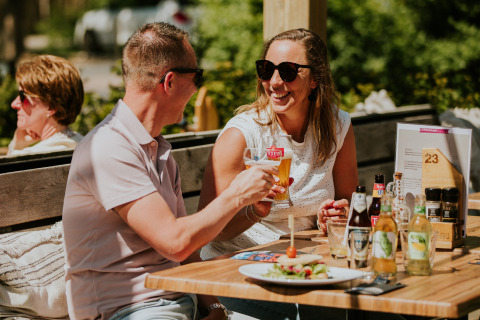 Dos personas disfrutan de bebidas y comida al aire libre en la terraza de un alojamiento glamping soleado.
