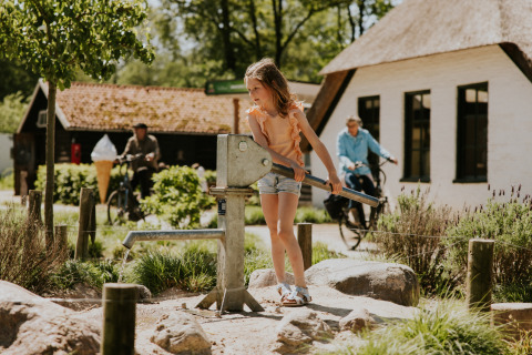 Una bambina pompa acqua a una pompa a mano fuori dalle cabine in un glamping, con ciclisti sullo sfondo.