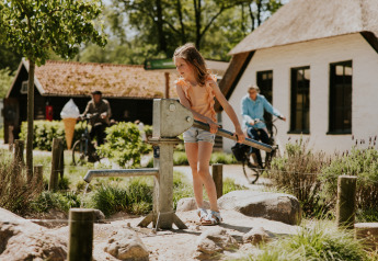 Ein Mädchen pumpt Wasser an einer Handpumpe vor Hütten auf einer Glamping-Unterkunft, Radfahrer im Hintergrund.
