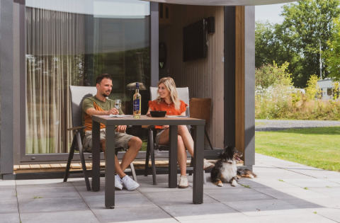 Couple enjoys wine outdoors at a table with their dog, outside a cosy tiny house at Campsite Si-Es-An.
