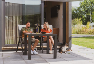Coppia beve vino all’aperto con il cane davanti a una tiny house al campeggio Si-Es-An nei Paesi Bassi.