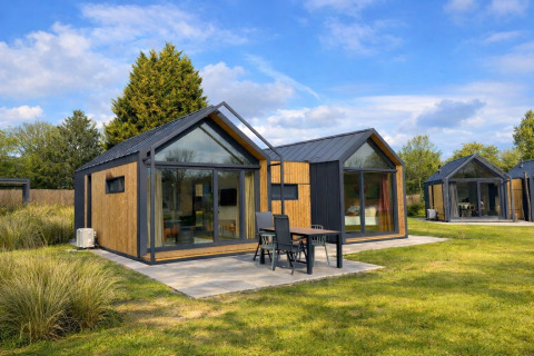 Modern Cosy Lodge with large glass windows, outdoor seating and green lawn under a blue sky.