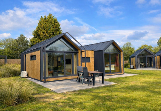 Moderne Cosy Lodge med store vinduer og udendørs siddeplads i grønne omgivelser under blå himmel.
