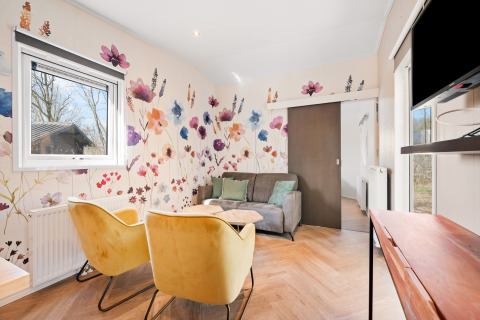 Zona giorno luminosa alla Happy bee lodge, Campeggio Si-Es-An, Paesi Bassi, con parete floreale e poltrone moderne.