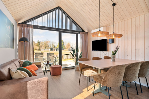 Moderne stue i Reestdal-lodgen på Si-Es-An campingplads i Nederlandene med store vinduer og spiseområde.