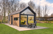 Modern lodge called Reestdal Loft with large windows and patio, set amidst green grass and surrounding trees.