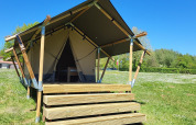 Safaritent met houten terras op Camping La Colline in België, gelegen op een groen grasveld onder blauwe lucht.