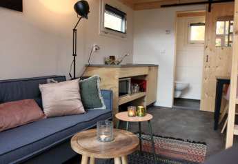 Gezellige woonkamer van een tiny house in De Hooge Veluwe met bank, kussens, tafeltjes en zicht op de badkamer.