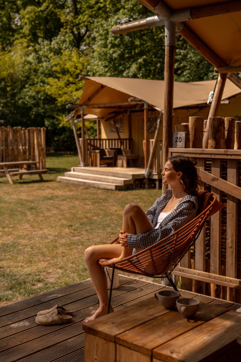 Donna rilassata sulla veranda della tenda safari De Bos Escape a Gooise Heide, Paesi Bassi, giornata di sole.