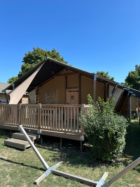 Safari tent called Villatent Outback on raised wooden deck, with greenery and clear blue sky above.