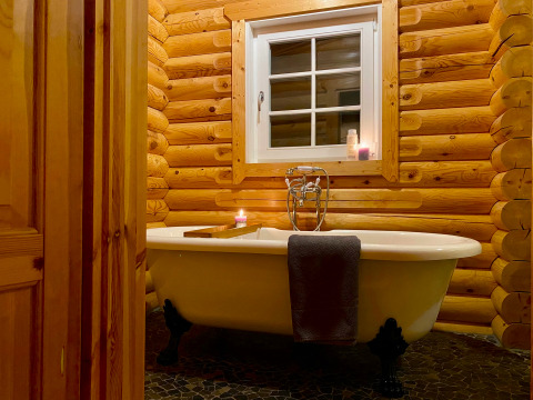 Cozy lodge bathroom with a freestanding clawfoot tub, candles, log walls, and a window above.