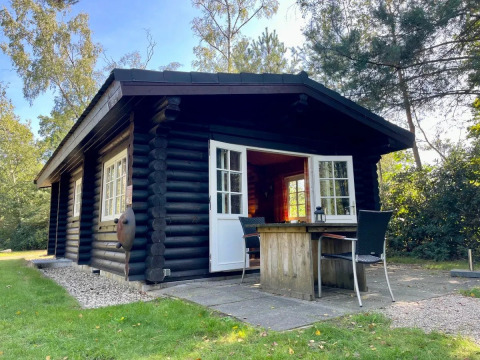 Cabaña de madera negra con ventanas y puertas blancas, terraza con mesa y sillas, rodeada de naturaleza.