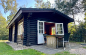 Cabaña de madera negra con ventanas y puertas blancas, terraza con mesa y sillas, rodeada de naturaleza.