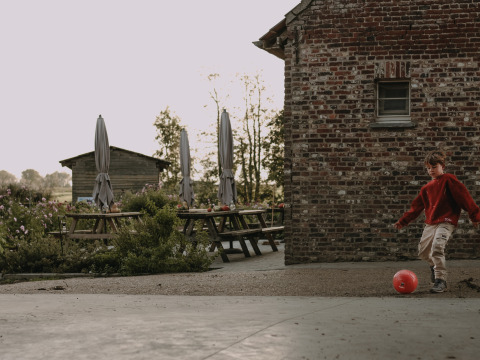 A boy in a red sweater kicks a red ball beside a brick building at BoerenBed Hoeve den Overdraght, Belgium.