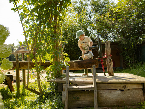 Un niño con gorra llena una tetera en una bomba de agua al aire libre en el jardín soleado de BoerenBed De Lange Weide, Drenthe.
