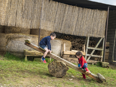 Due bambini giocano su un'altalena di legno in un ambiente rurale con balle di fieno e legna sullo sfondo.