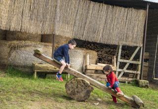 Due bambini giocano su un'altalena di legno in un ambiente rurale con balle di fieno e legna sullo sfondo.