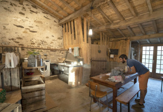 Uomo che lavora a un tavolo di legno in una cucina rustica con muri in pietra e travi in legno a vista.