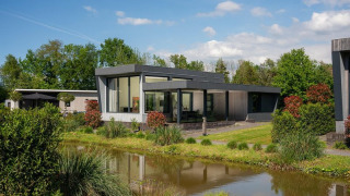 Moderne Boslodge Plus med hottub, spejlet i en dam, omgivet af frodigt grønt og blå himmel.