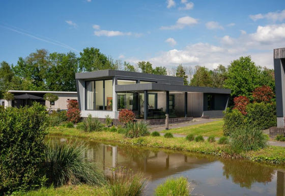 Moderne Boslodge Plus med hottub, spejlet i en dam, omgivet af frodigt grønt og blå himmel.