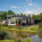 Moderne Boslodge Plus met hottub aan water, omgeven door groen, bomen en een blauwe lucht.
