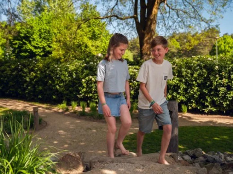 To børn går barfodet på en sti i ferieparken Molenvelden, omgivet af grønne træer og buske.