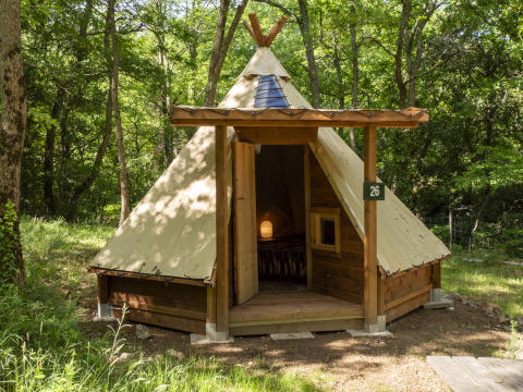 Modern tipi tent in the woods with wooden structure and an open door revealing a cozy, lit interior.