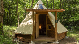 Modern tipi tent in the woods with wooden structure and an open door revealing a cozy, lit interior.