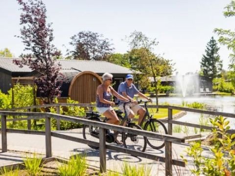 Two adults cycling in Holiday Park Molenvelden, North Brabant, Netherlands, past a pond and fountain.