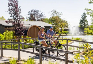 To voksne cykler gennem ferielandet Molenvelden i Nord-Brabant, Holland, med søen og springvand bagved.