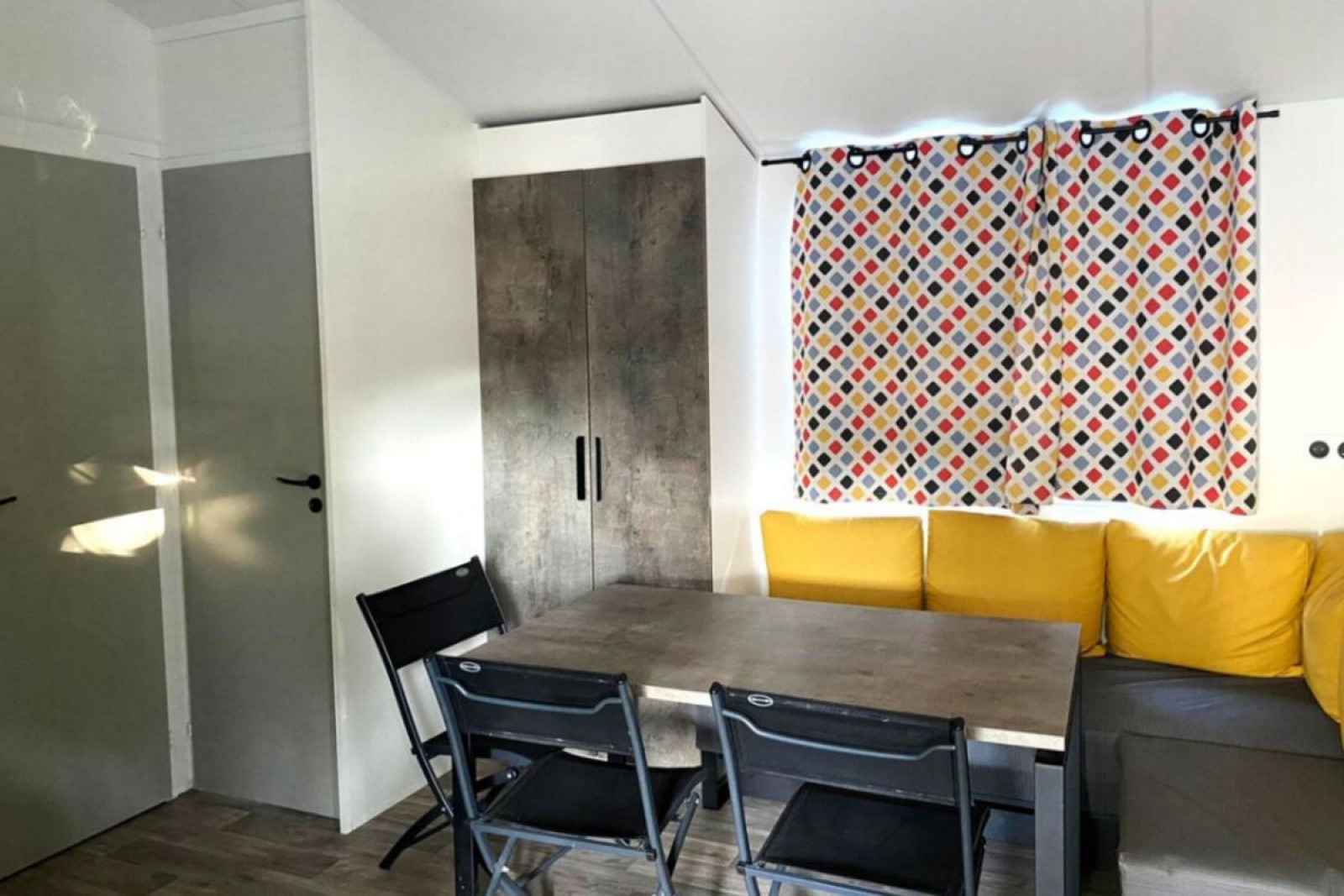 Eethoek met grijze tafel, zwarte stoelen, gele kussens en kleurrijk gordijn in Comfort Mobile home Frankrijk.