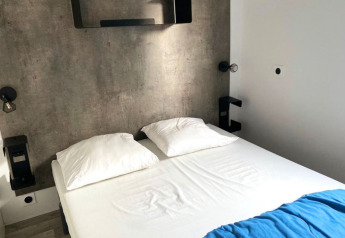 Modernes Glampingzimmer mit Betonwand, einem Doppelbett mit weißen Kissen und blauer Bettdecke.