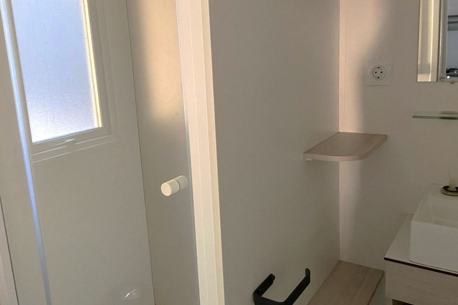 Kleine badkamer met witte deur, wastafel, spiegel en zwarte wc-rolhouder in een Frans mobiel huis.